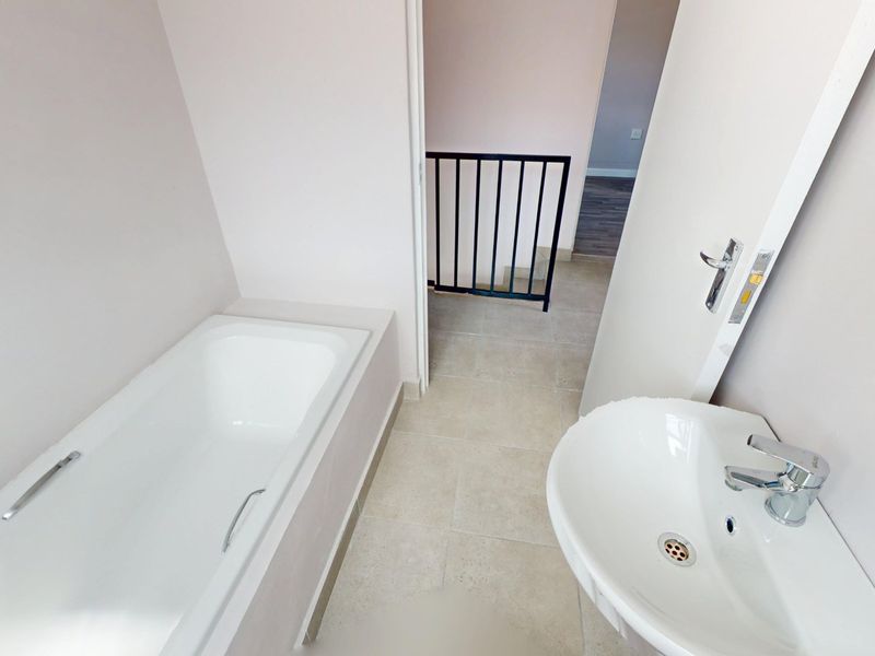 A clean, modern bathroom featuring a built-in bathtub and pedestal washbasin, with neutral tiled flooring and direct access to the staircase landing.
