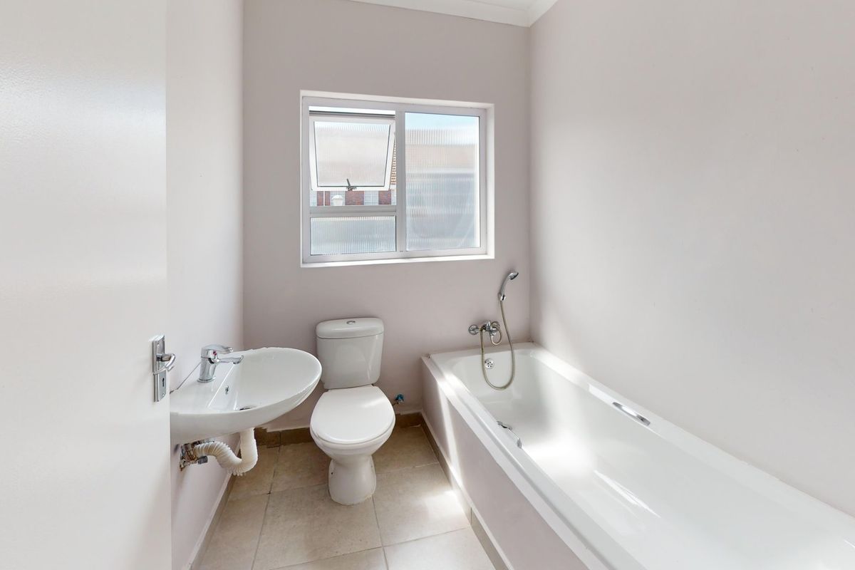 A clean, functional bathroom featuring a white bathtub, pedestal basin, and toilet with neutral tiled flooring and natural light from a sliding window.