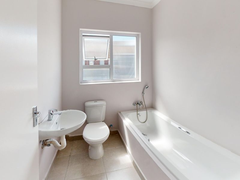A clean, functional bathroom featuring a white bathtub, pedestal basin, and toilet with neutral tiled flooring and natural light from a sliding window.