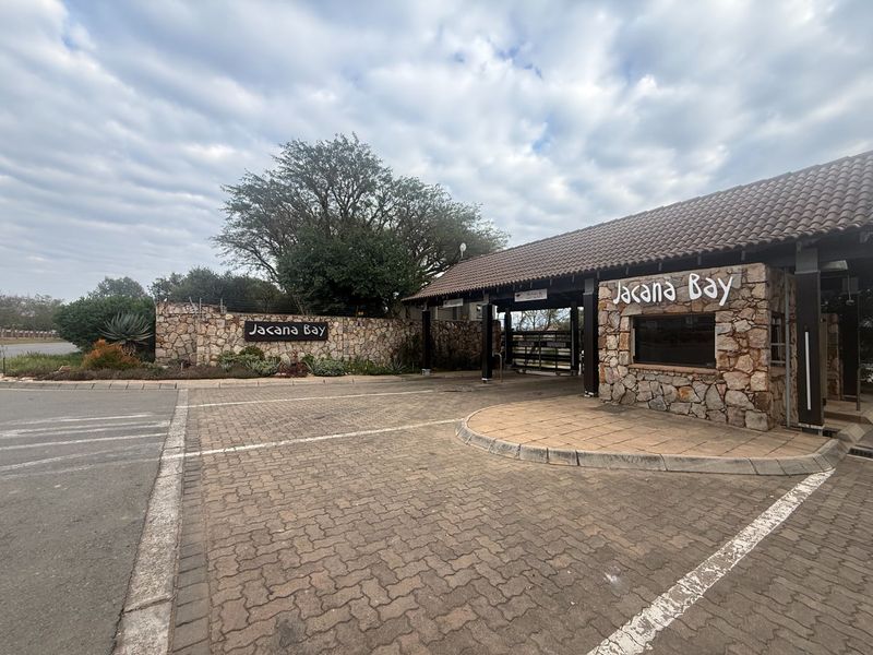 The entrance to Jacana Bay estate featuring a stone-clad security guardhouse, paved driveway, and gated access point.
