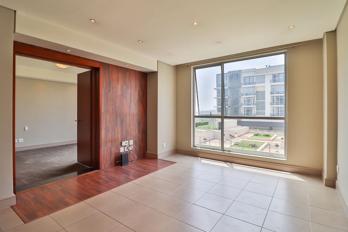 An empty, well-lit living area featuring tiled floors, a timber accent wall, and a large window with views of the surrounding apartment complex.