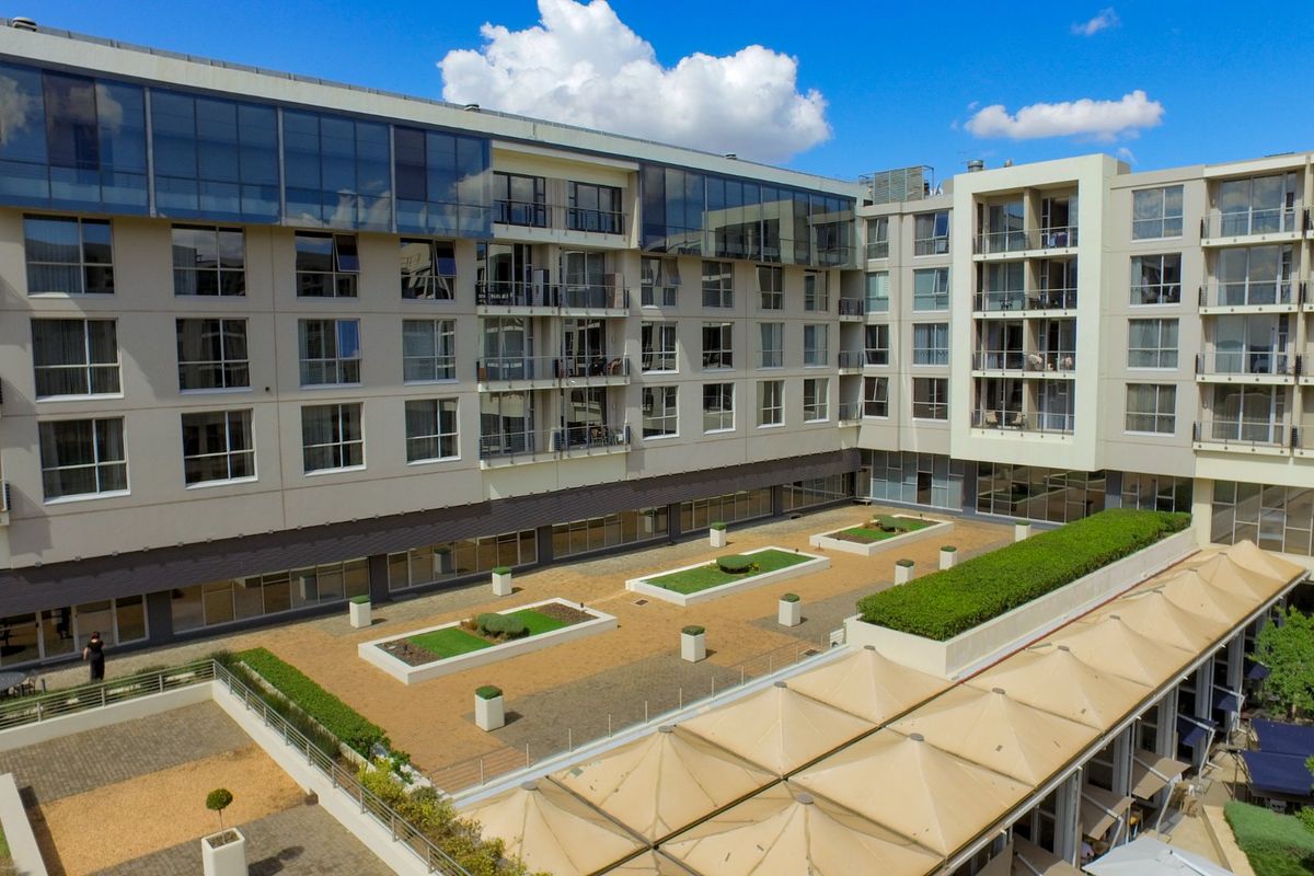 An aerial view of a modern apartment complex featuring a central paved courtyard, landscaped garden beds, and multi-level balconies with glass railings.