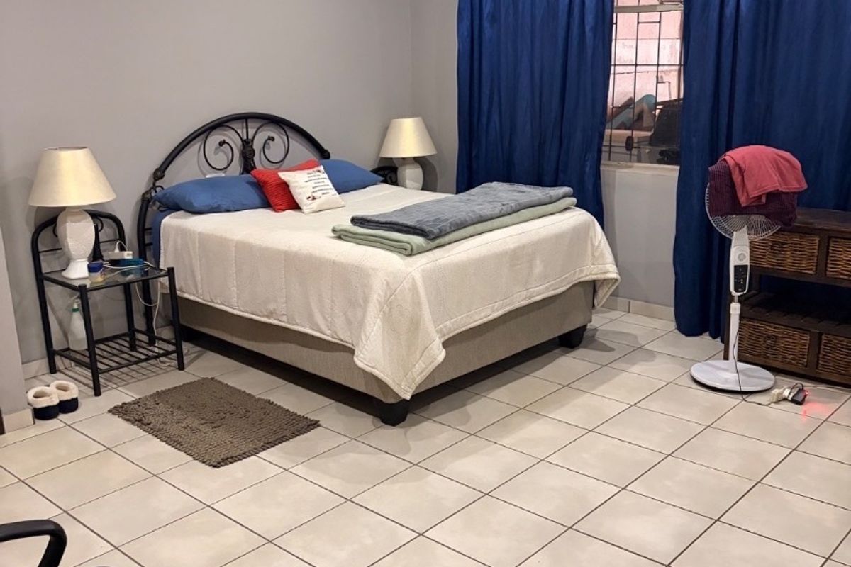 A spacious bedroom featuring neutral tiled flooring, a ceiling fan, and security bars on the window, offering a functional and well-lit living space.
