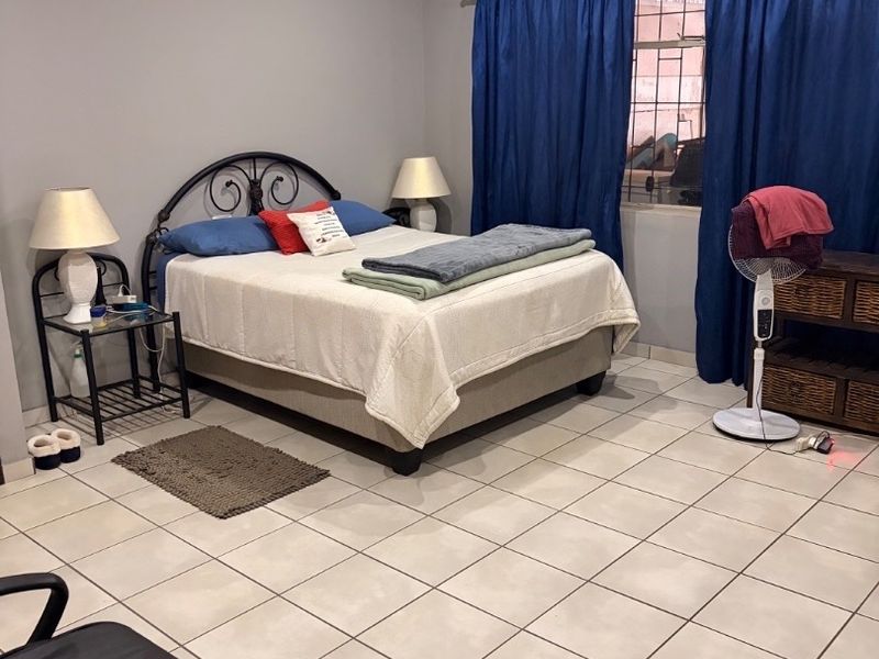 A spacious bedroom featuring neutral tiled flooring, a ceiling fan, and security bars on the window, offering a functional and well-lit living space.
