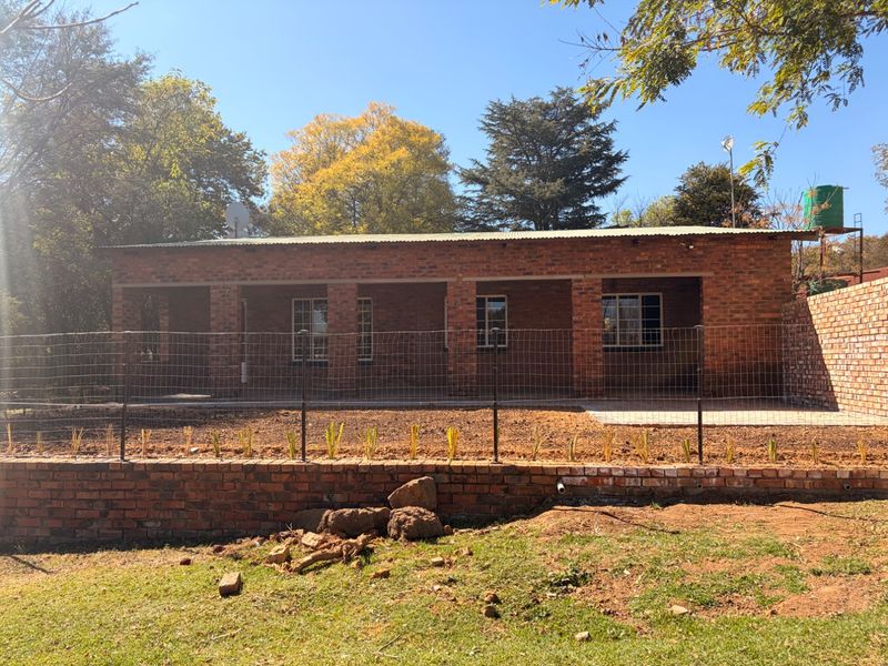 A rear view of a single-story exposed brick house featuring a covered patio, wire fencing, and a brick retaining wall in a garden setting.