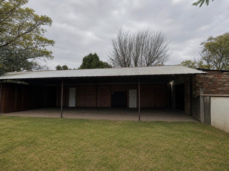 A spacious rear outdoor area featuring a large covered patio with brick walls and a corrugated metal roof, overlooking a grass lawn.