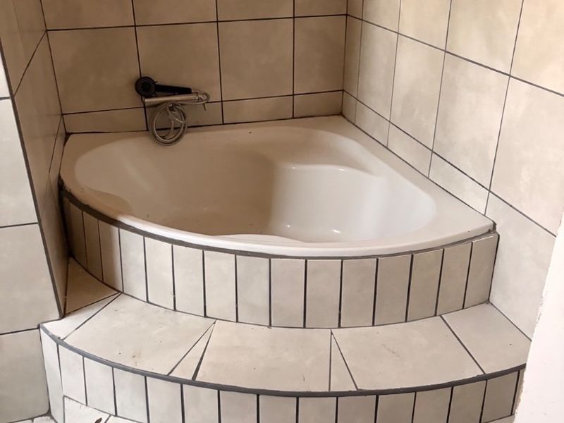 A corner bathtub featuring a raised tiled platform and steps, requiring some maintenance to the grout and finishes.
