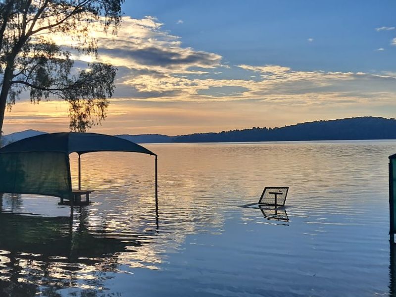 A scenic view of a lake at sunset, showing significant flooding affecting the shoreline and outdoor structures.