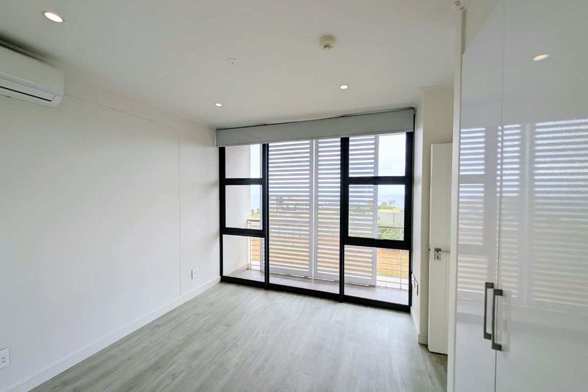 A bright, empty bedroom featuring modern laminate flooring, built-in cupboards, a split-system air conditioner, and large sliding doors leading to a balcony.
