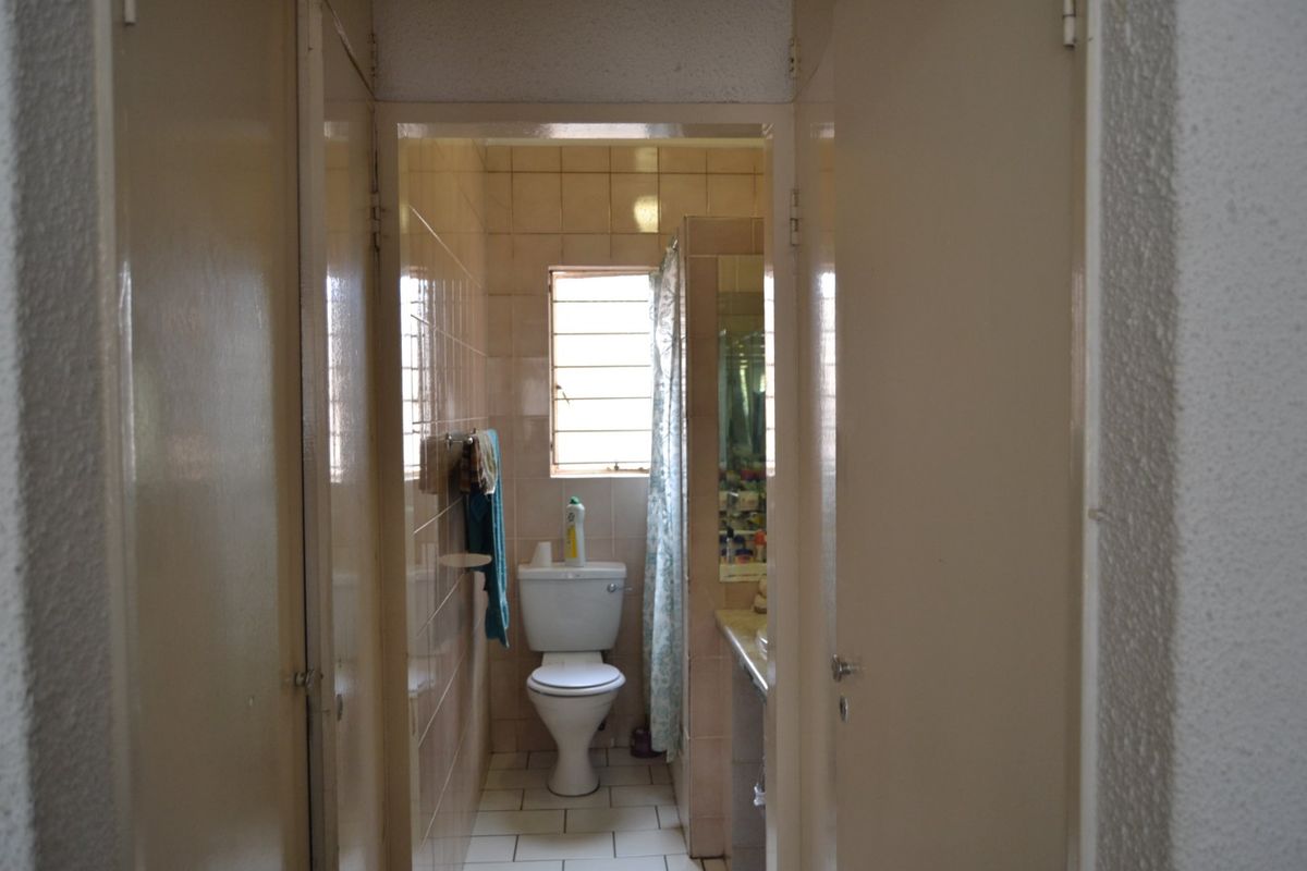 A dated bathroom featuring ceramic tiling, a standard toilet, and a vanity basin, accessible through a narrow hallway with built-in storage doors.