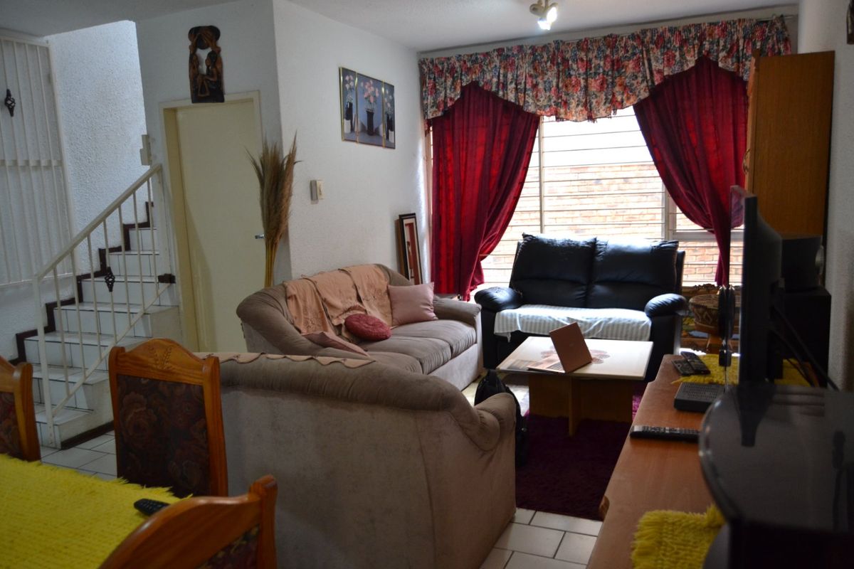 A living room area featuring tiled flooring, a staircase with a security gate, and large windows fitted with security bars.
