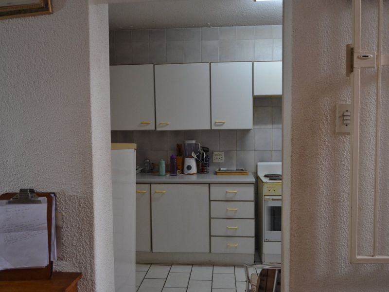 A compact, dated kitchen featuring white cabinetry, tiled backsplash, and tiled flooring, viewed through a doorway.
