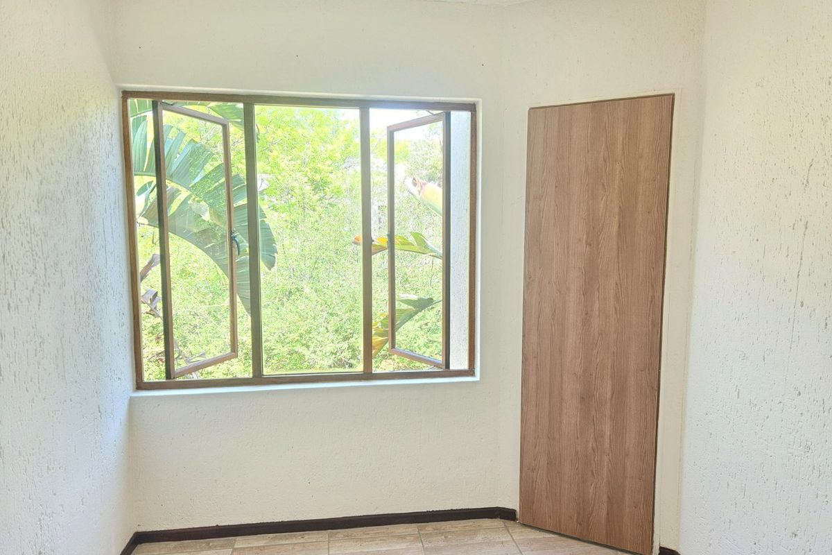 A bright, empty bedroom featuring tiled flooring, a wood-finish door, and a large window overlooking lush greenery.