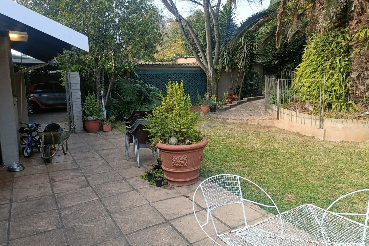 A rear garden area featuring a paved patio, established trees, and a metal fence, providing a functional outdoor space for relaxation.