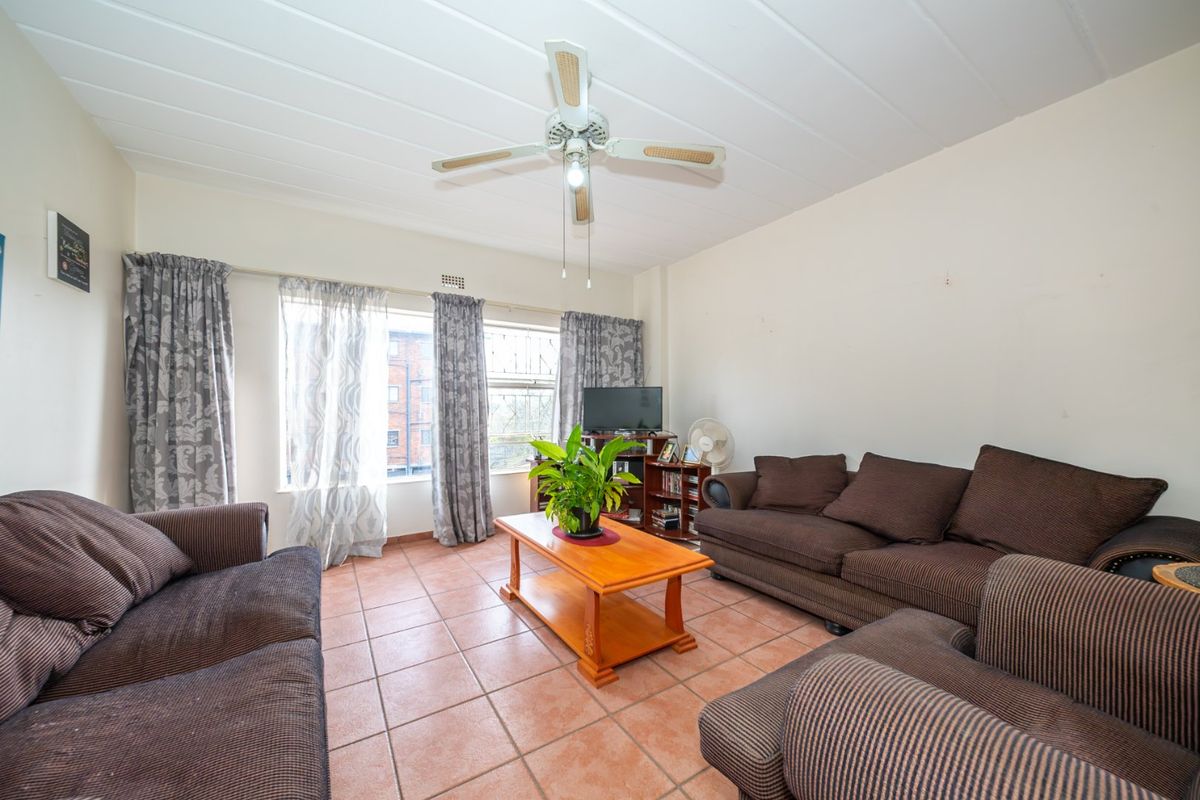 A spacious living room featuring tiled flooring, a ceiling fan, and large windows that provide natural light.