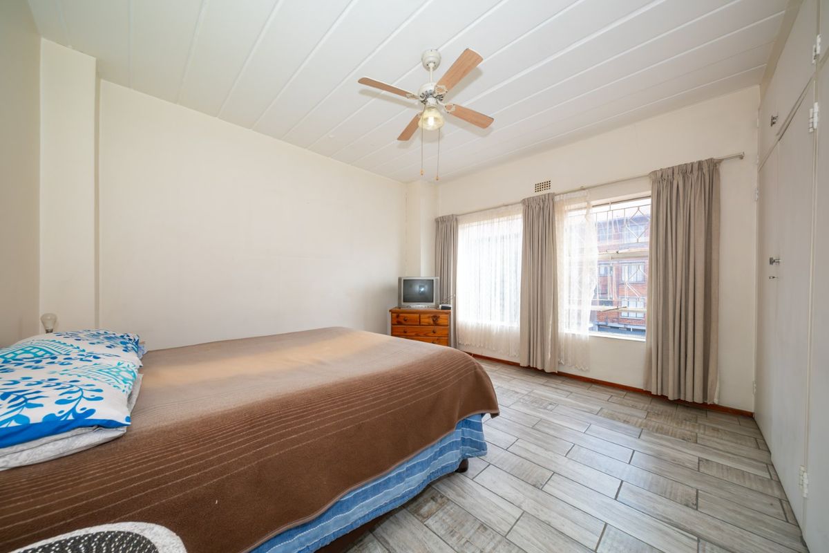 A bedroom featuring tiled flooring, a ceiling fan, and built-in cupboards, with natural light provided by large windows fitted with security bars.