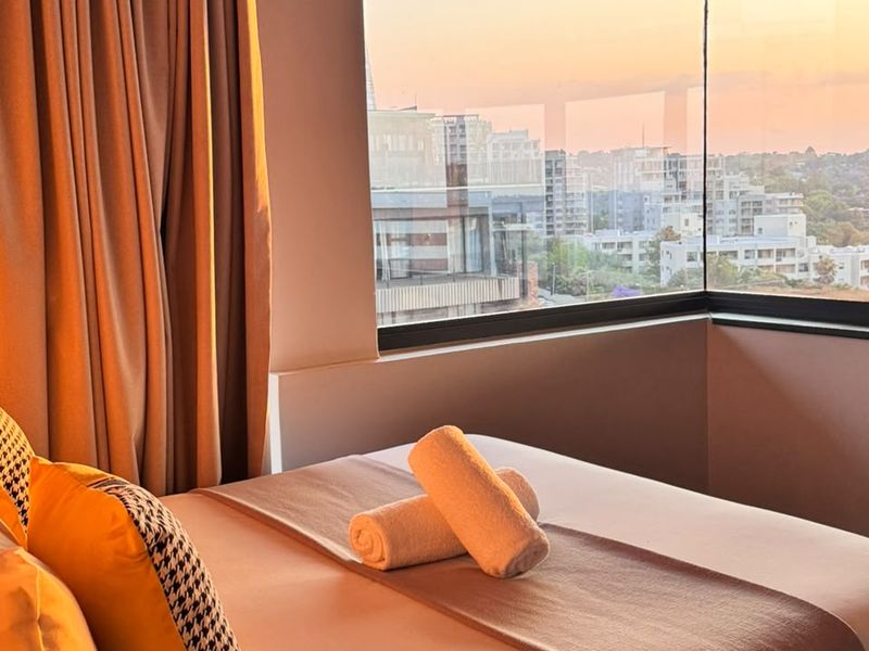 A well-lit bedroom featuring a large corner window that offers expansive city views and natural light.