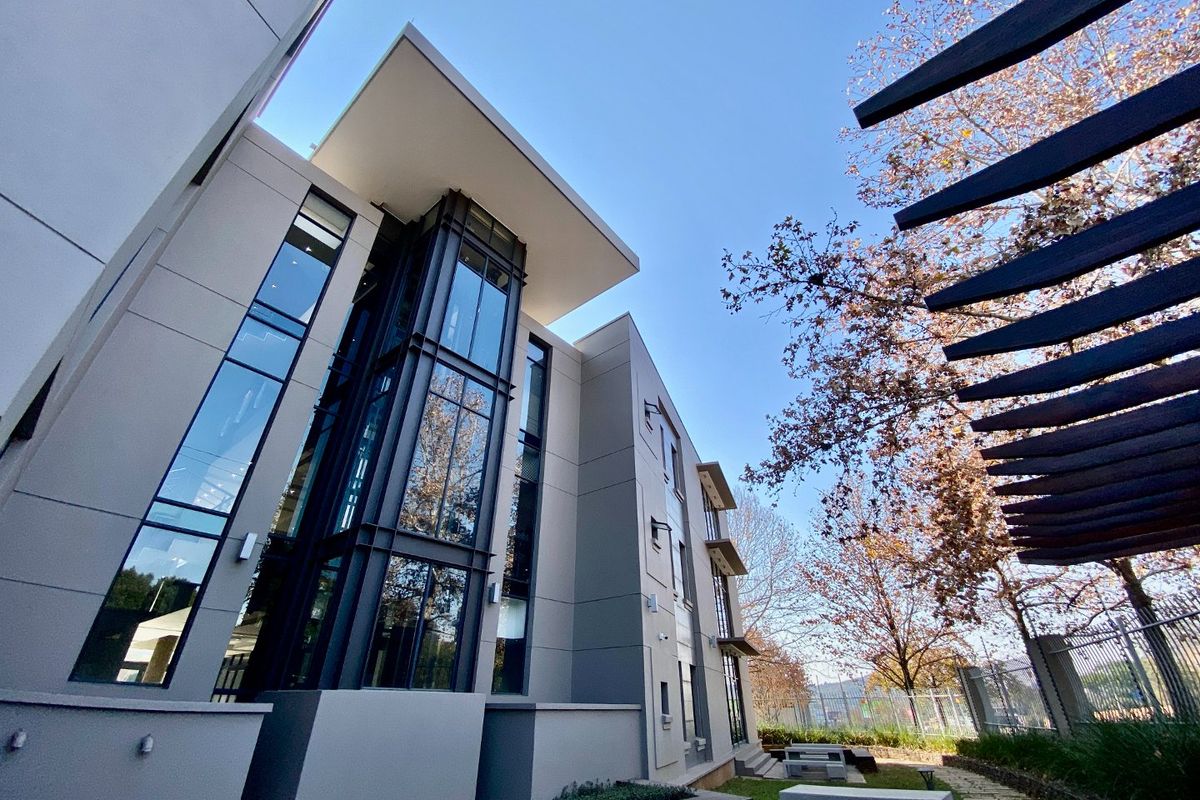 Modern commercial building exterior featuring large glass curtain walls, steel structural elements, and a contemporary flat roof design with adjacent landscaped outdoor area.
