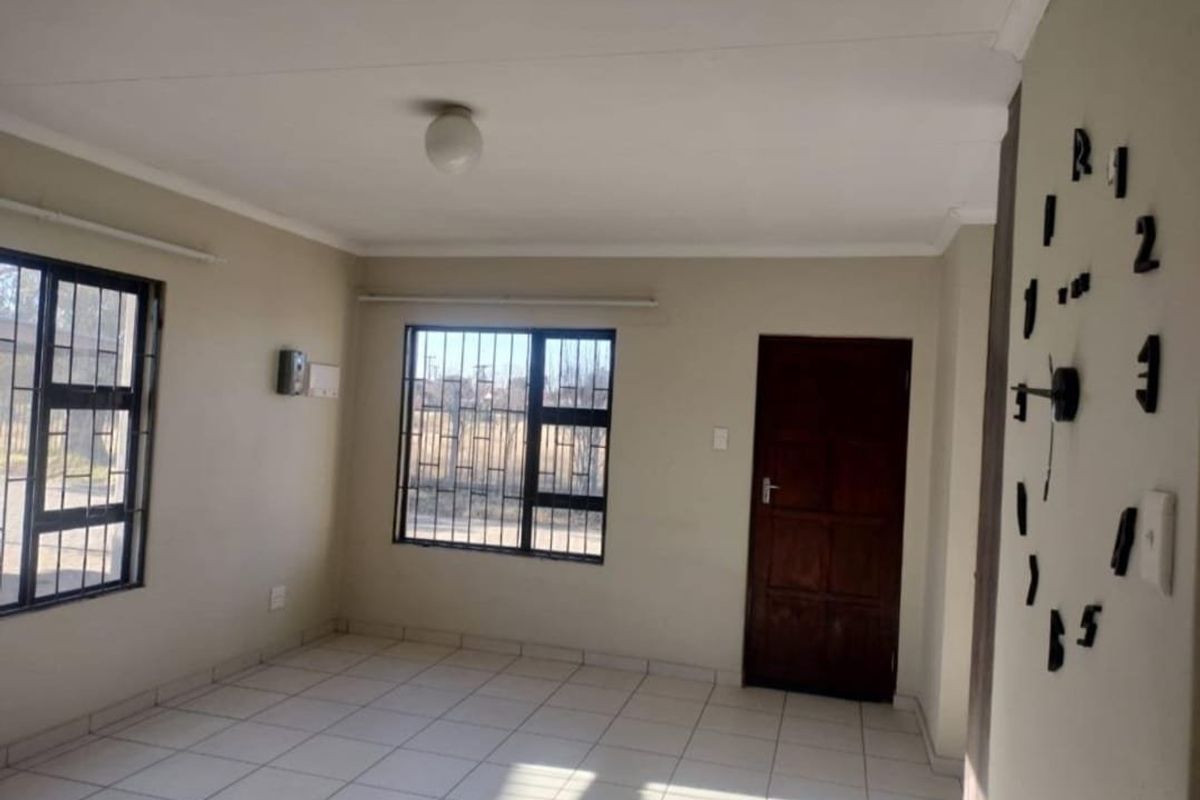 A bright, empty living room featuring neutral tiled flooring, security bars on the windows, and a solid timber door.