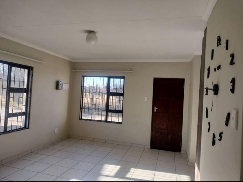 A bright, empty living room featuring neutral tiled flooring, security bars on the windows, and a solid timber door.