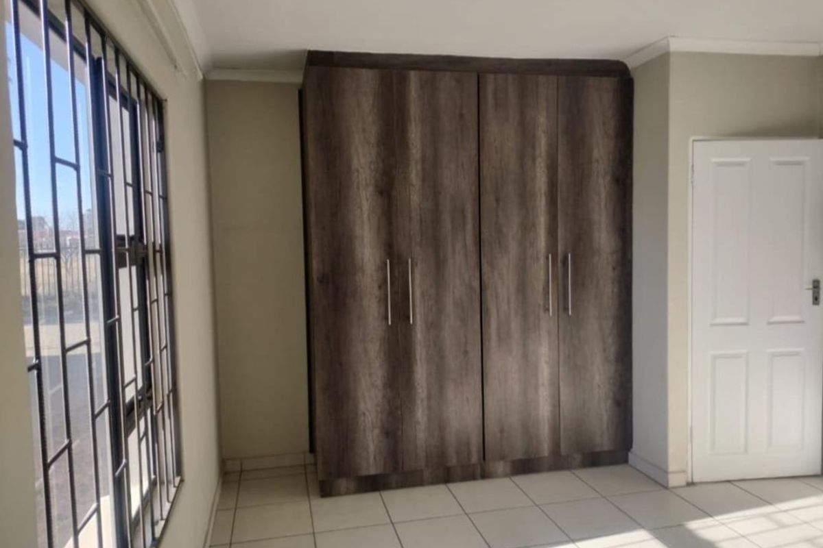 A bright, empty bedroom featuring modern wood-finish built-in cupboards, neutral tiled flooring, and security bars on the window.