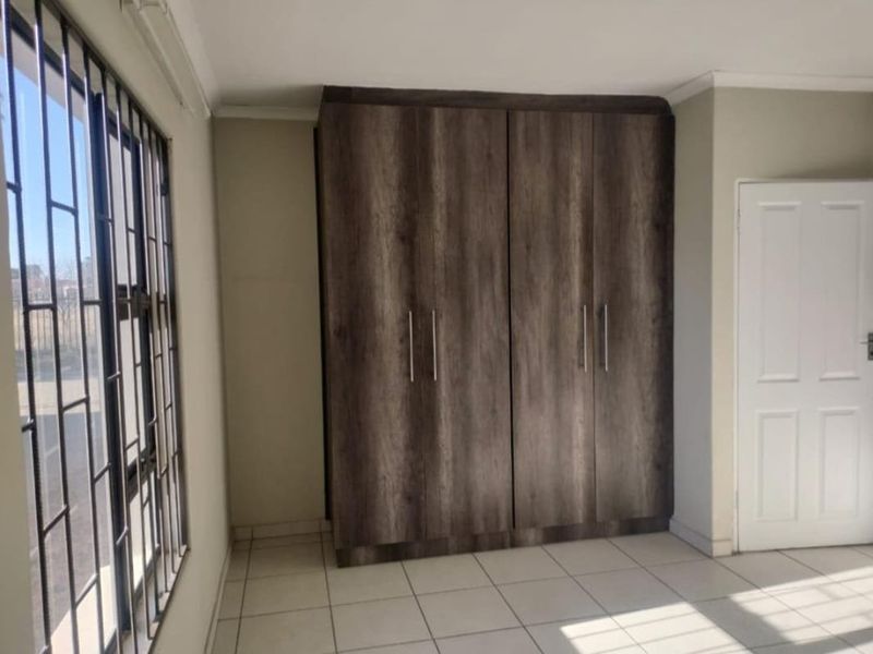 A bright, empty bedroom featuring modern wood-finish built-in cupboards, neutral tiled flooring, and security bars on the window.