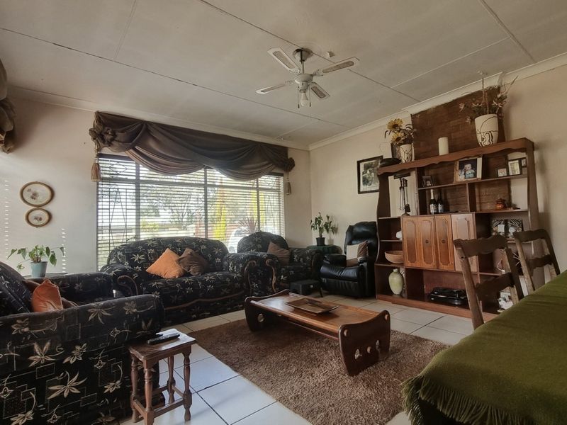 A living room featuring tiled flooring, a ceiling fan, and a brick feature wall, with visible wear on the ceiling panels.