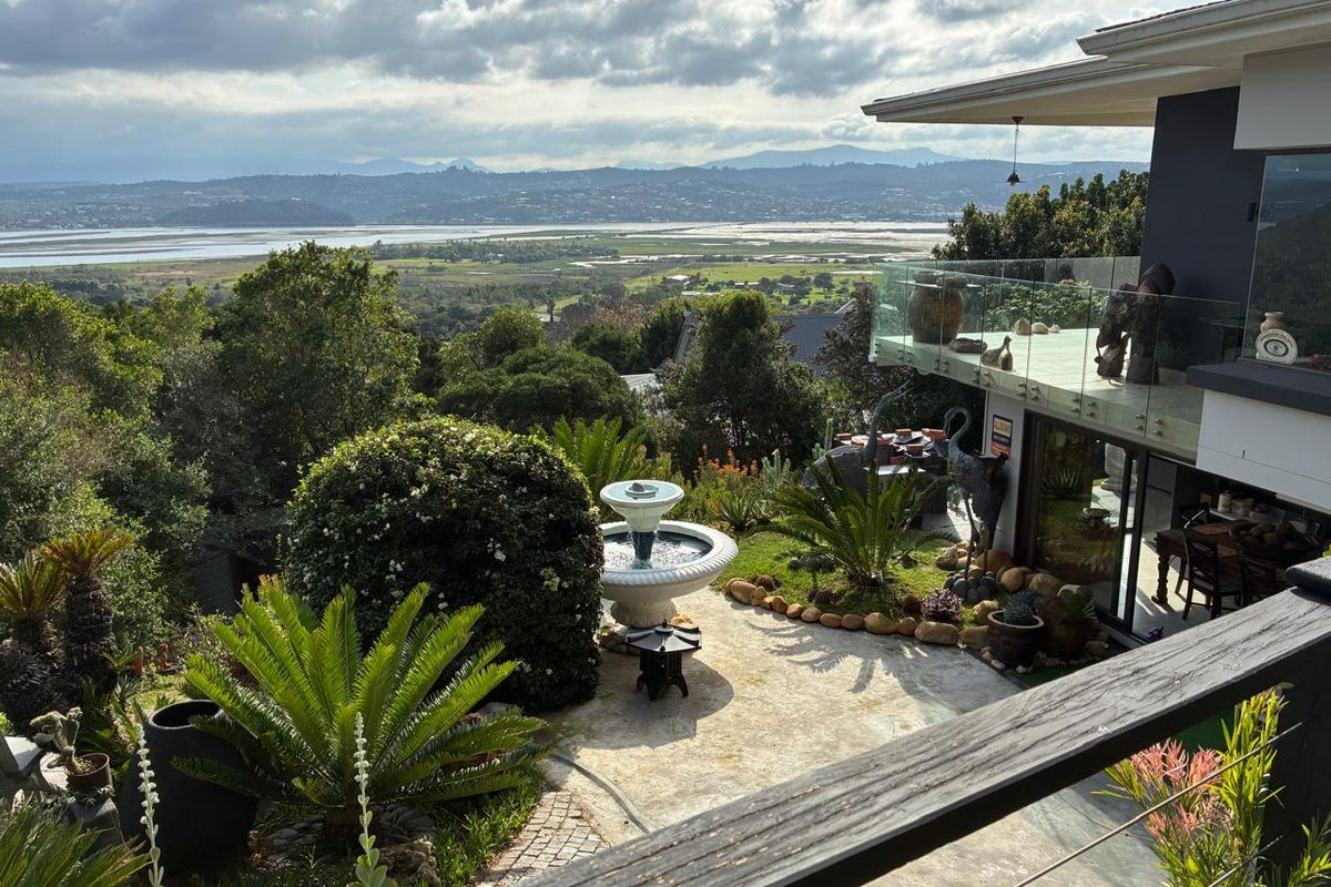 Rear exterior view featuring a landscaped garden, stone patio with a water fountain, and a modern balcony with a glass balustrade overlooking the landscape.