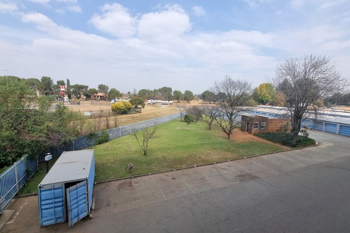 An elevated view of the property grounds featuring a paved driveway, landscaped lawn, perimeter fencing, and a row of storage units.