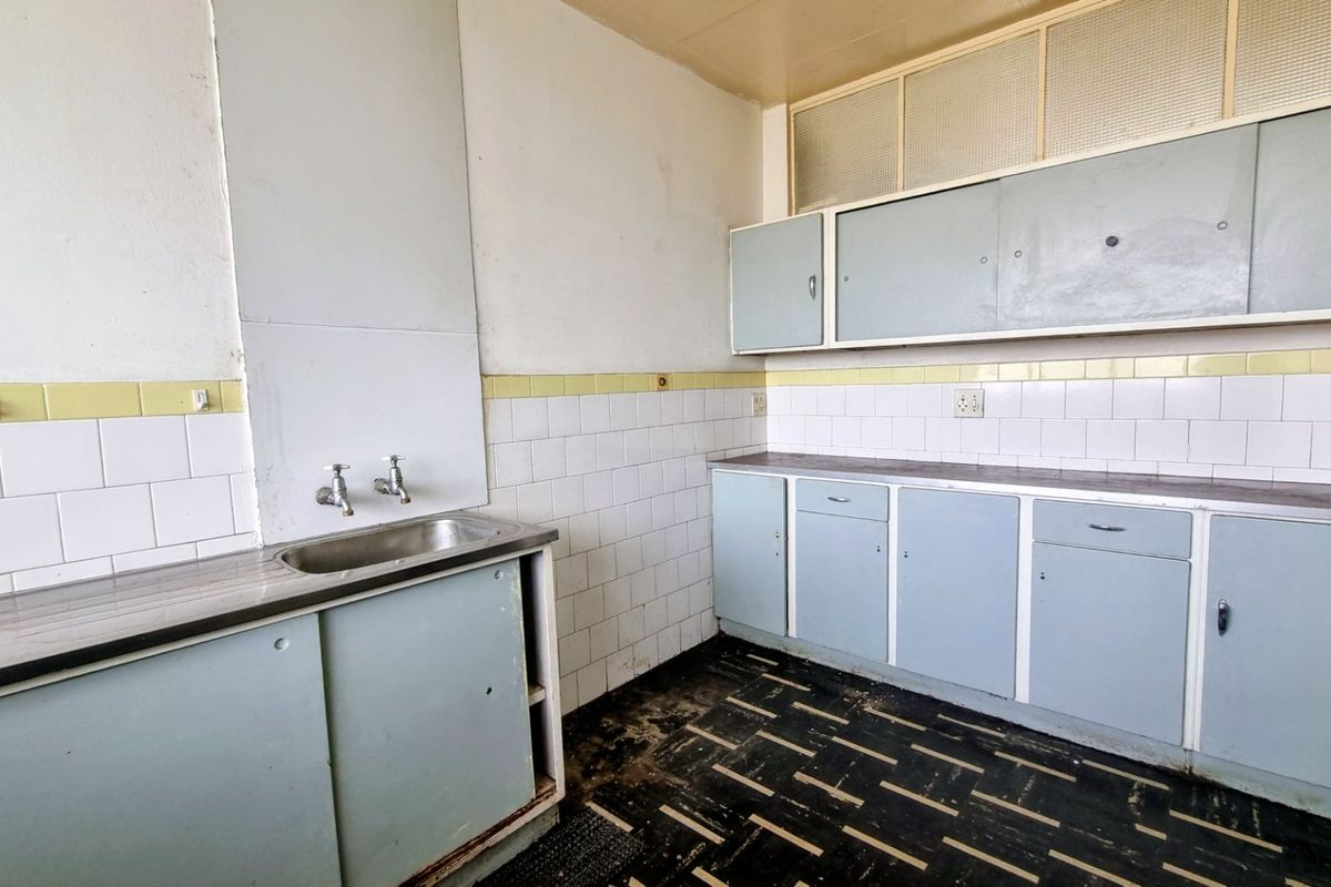 A dated, empty kitchen featuring original built-in cabinetry, tiled backsplash, and a stainless steel sink, requiring renovation to modernize the space.