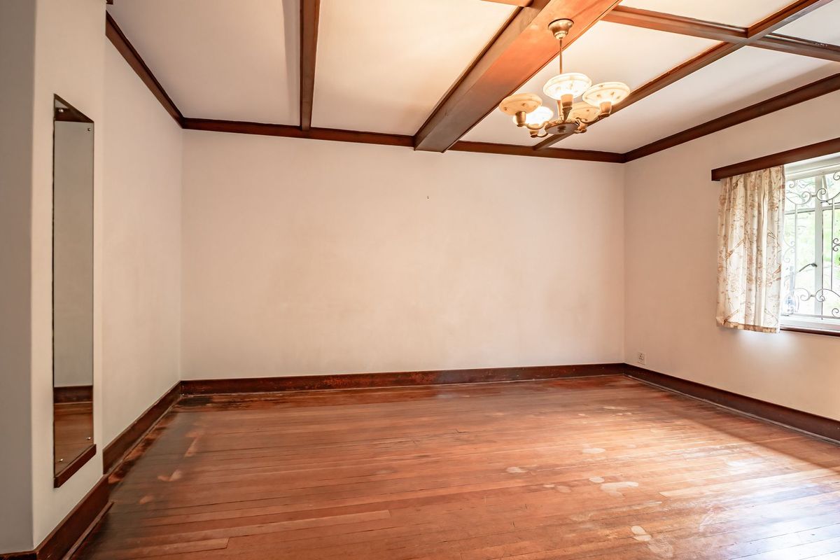 An empty bedroom featuring original hardwood flooring, exposed timber ceiling beams, and a window fitted with security bars.