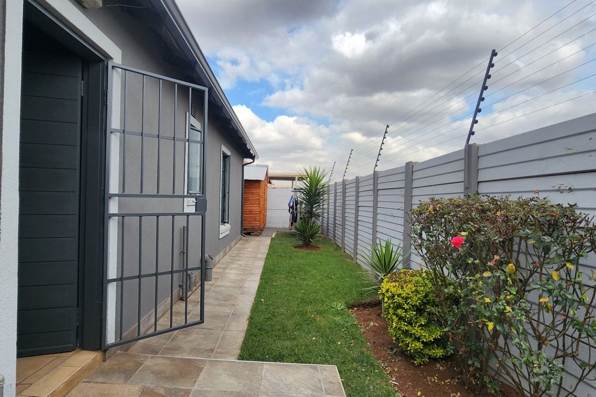 A side garden area featuring a tiled walkway, secure boundary wall with electric fencing, and a security gate leading to the home.
