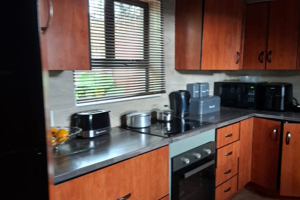 A functional kitchen featuring warm-toned wooden cabinetry, dark laminate countertops, an integrated oven and hob, and classic tiled flooring with exposed ceiling beams.