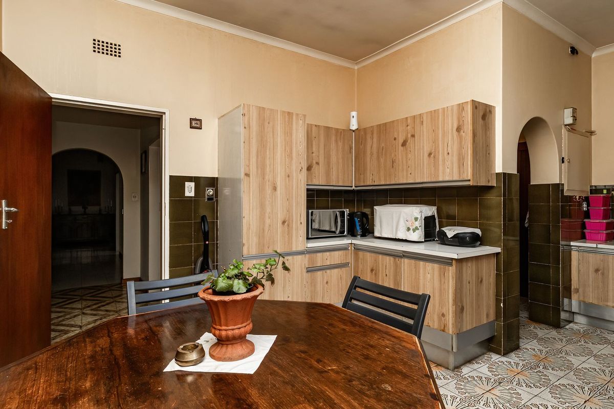 A dated kitchen featuring wood-look cabinetry, dark tiled splashbacks, and patterned floor tiles, with an open archway leading to adjacent living spaces.