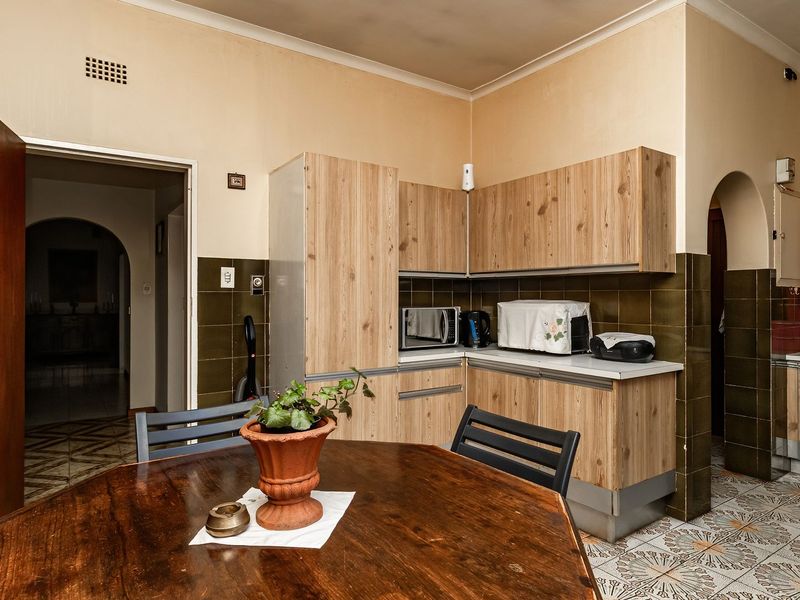 A dated kitchen featuring wood-look cabinetry, dark tiled splashbacks, and patterned floor tiles, with an open archway leading to adjacent living spaces.