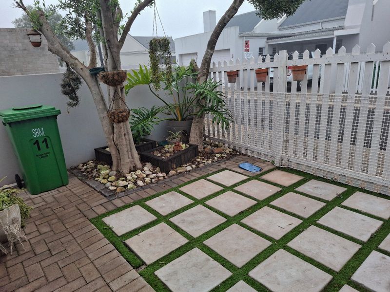 A compact, well-maintained garden area featuring a paved patio, artificial grass accents, and a white picket fence boundary.