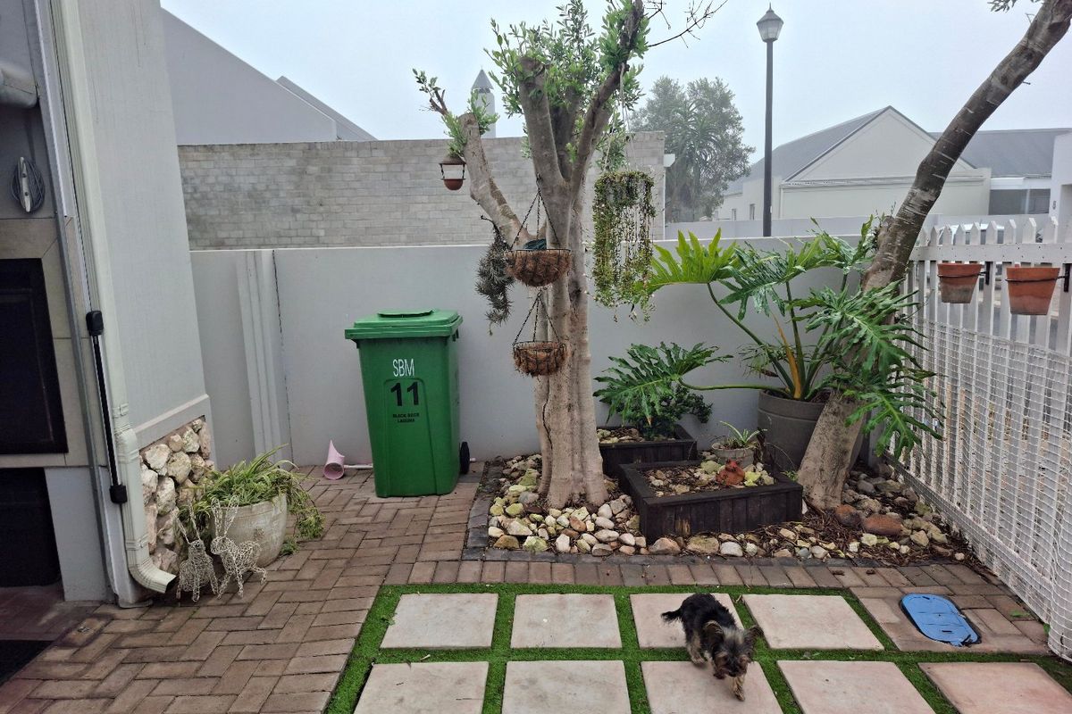 A compact rear courtyard featuring paved areas, artificial lawn with stepping stones, a built-in braai, and established garden beds.