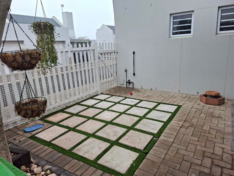 A compact rear courtyard featuring a paved patio area with artificial grass inlays, enclosed by a white picket fence.