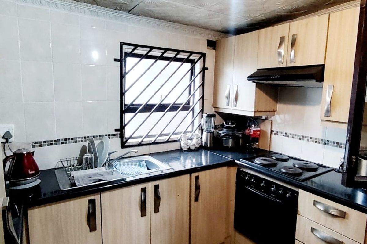 Compact kitchen featuring light wood-finish cabinetry, black countertops, built-in oven and hob, and tiled backsplash with security bars on the window.