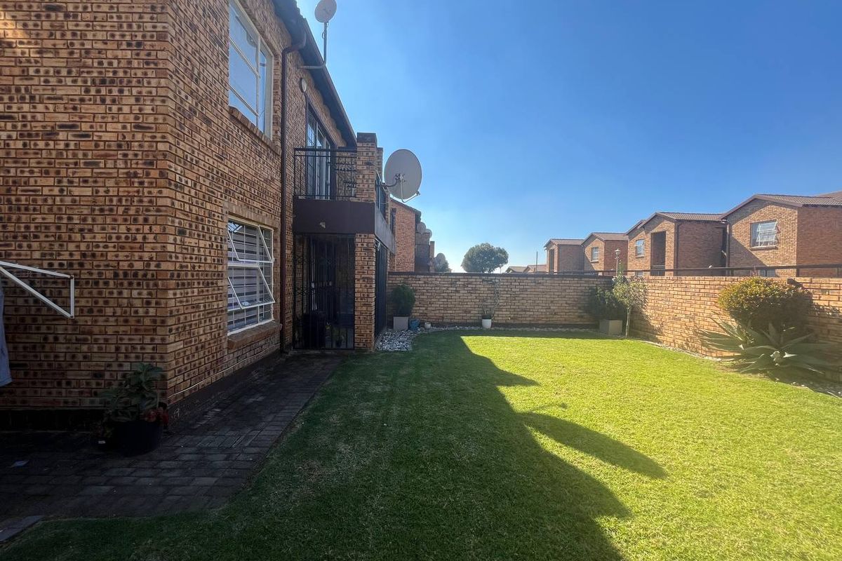 Rear view of the townhouse featuring a face brick facade, a small balcony, a paved walkway, and a private lawn area enclosed by boundary walls.