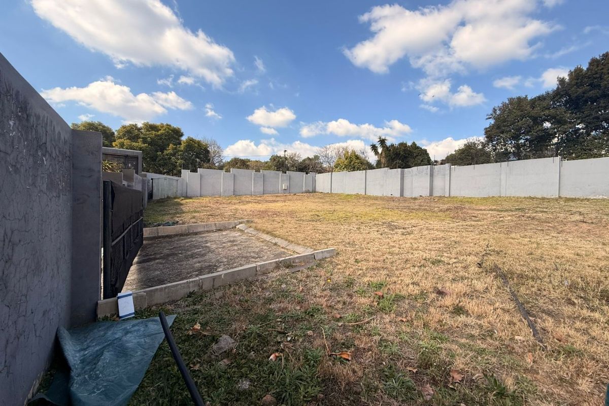 A vacant residential plot featuring a perimeter wall, a small concrete slab, and a metal gate, offering a clear space for development.