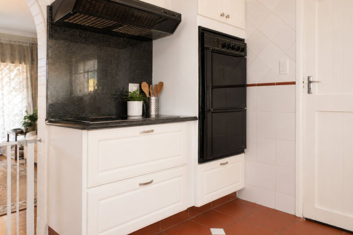 A functional kitchen area featuring white shaker-style cabinetry, a black granite countertop, built-in oven, and extractor fan with terracotta-style floor tiling.