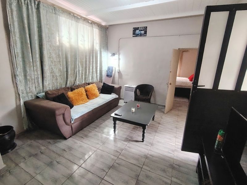 A living room featuring tiled flooring and exposed ceiling beams, with an adjacent bedroom visible through the doorway.