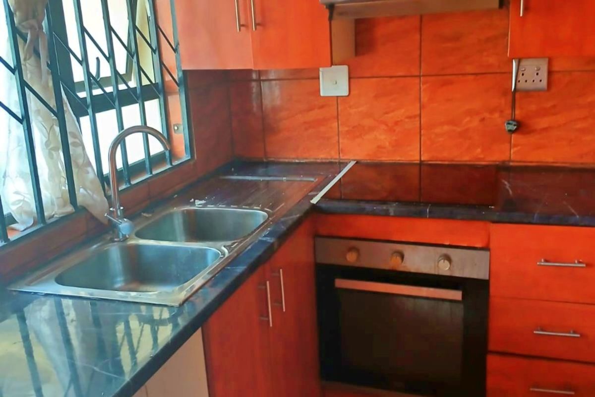 Compact kitchen featuring orange-toned cabinetry, dark countertops, a built-in oven, hob, and extractor fan, with security bars on the window.