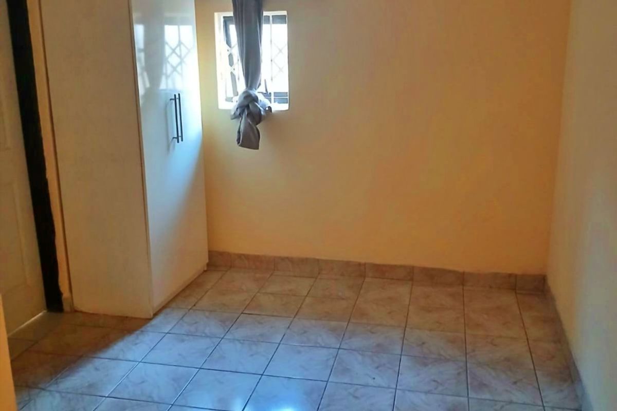A simple, empty bedroom featuring tiled flooring, a ceiling fan, and a built-in cupboard with security bars on the window.
