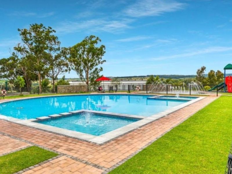 A well-maintained outdoor swimming pool area featuring a paved deck, water fountains, and surrounding landscaped lawn with safety fencing.