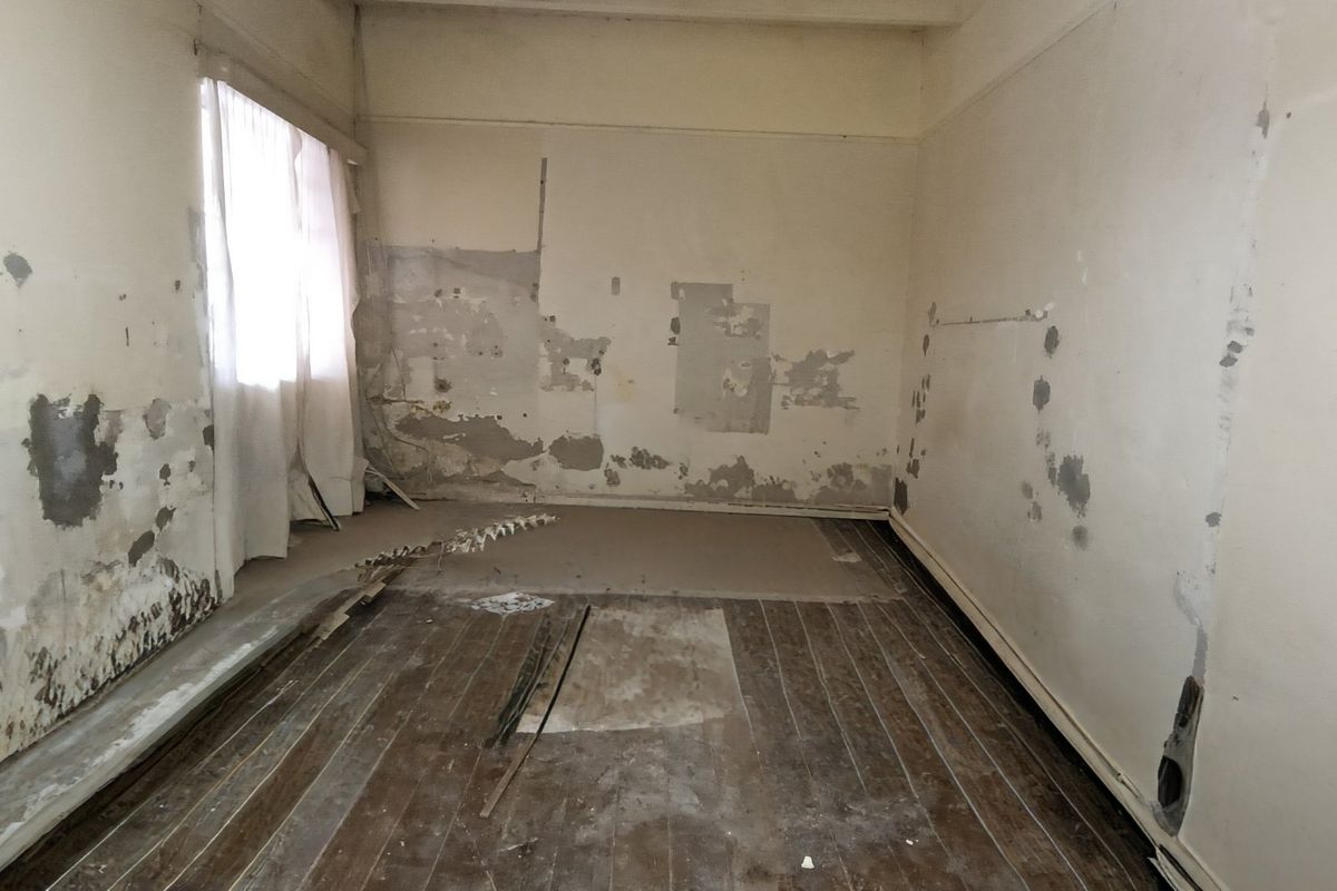 An empty bedroom requiring significant renovation, featuring original timber flooring and damaged wall surfaces in need of repair and refurbishment.