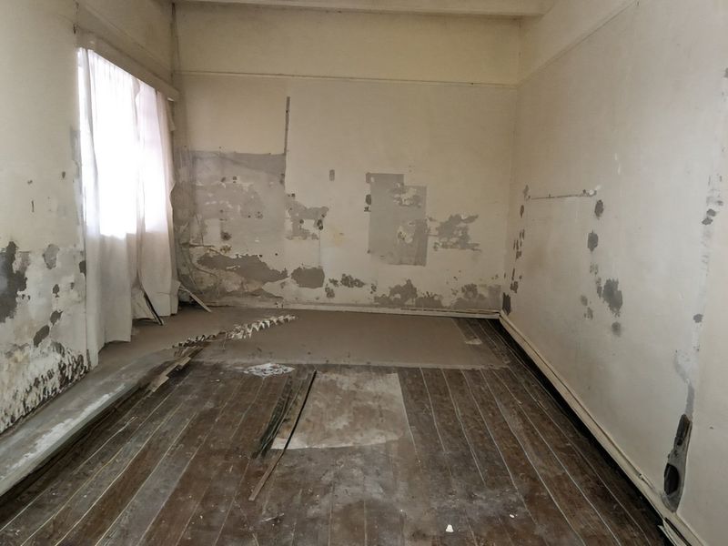 An empty bedroom requiring significant renovation, featuring original timber flooring and damaged wall surfaces in need of repair and refurbishment.