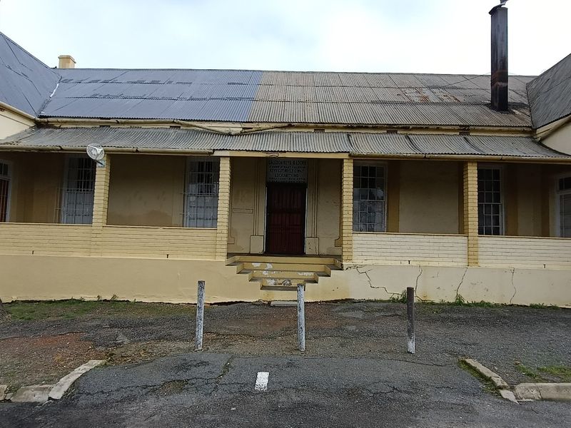 Front elevation of a single-story building featuring a covered veranda, corrugated iron roof, and visible masonry cracks requiring maintenance.