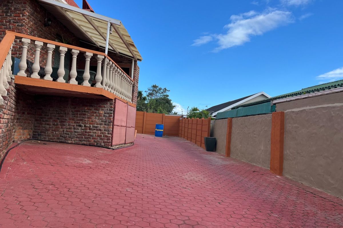 Rear exterior area featuring a brick facade, elevated balustrade balcony, paved driveway, and perimeter boundary walls.
